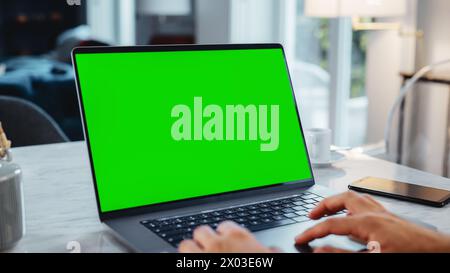 Green Screen Mock Up Display auf einem Laptop-Computer. Nahaufnahme der Hände von Personen, die von Modern Home aus arbeiten, das Touchpad verwenden und Inhalte scrollen. Das Smartphone liegt neben dem Computer. Stockfoto