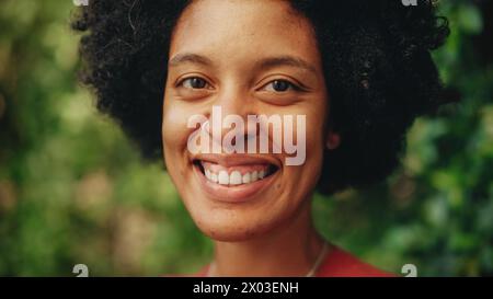Nahaufnahme Porträt einer glücklichen jungen Latina mit braunen Augen, Afrohaar und Nase Piercing für die Kamera. Schöne vielfältige multiethnische Schwarze Hispanic weibliche lächelnd auf grünem Natur Hintergrund. Stockfoto