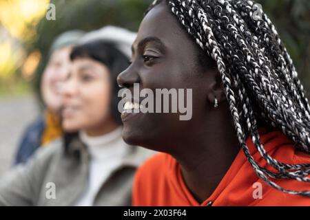 Leuchtende afrikanische Frau mit aufwändig geflochtenen Haaren lächelt herzlich - sanft verschwommener Hintergrund mit zwei jungen Frauen unterschiedlicher Ethnien Stockfoto