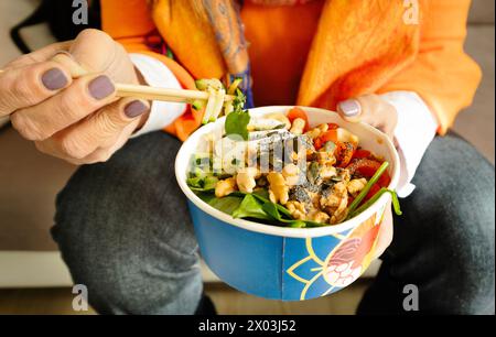 Nahaufnahme einer Frauenhand, die eine bunte Schüssel asiatischen Salats (Poke) mit Stäbchen hält Stockfoto