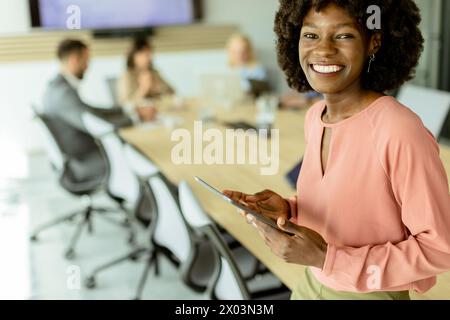 Ccheerful Afroamerikanerin leitet ein Meeting und hält sich an einem digitalen Tablet fest, während Kollegen hinter ihr Gespräche führen. Stockfoto