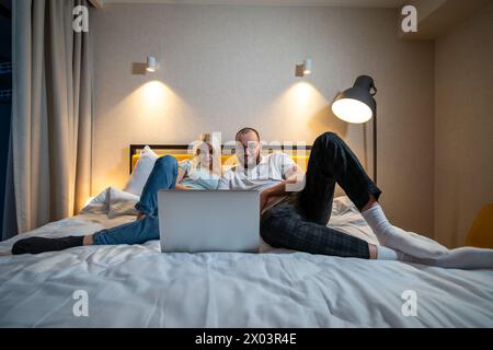 Ein Paar liegt auf dem Bett und teilt sich den Moment der Verbindung über den Laptop-Bildschirm in gemütlicher Schlafzimmeratmosphäre. Stockfoto