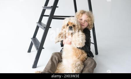 Ein kleiner Junge mit lockigen blonden Haaren sitzt auf dem Boden und lehnt sich an eine schwarze Stufenleiter, während er sich eng an sein großes, flauschiges, hellbraunes und anschmiegt Stockfoto