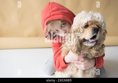 Ein glückliches Kind in einem roten Sweatshirt mit Kapuze auf dem Kopf spielt und umarmt einen Cocker Spaniel Hund Stockfoto