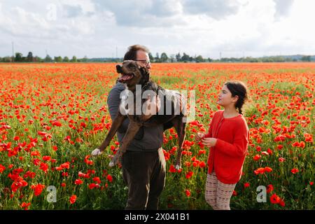 Porträt eines glücklichen Vaters, der einen großen Hund in den Armen hält und seine Tochter neben ihnen auf Mohnfeld steht Stockfoto