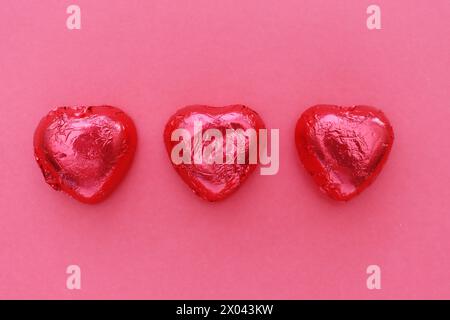 Drei herzförmige Pralinen in roter Folie auf rotem Hintergrund, Draufsicht. Stockfoto