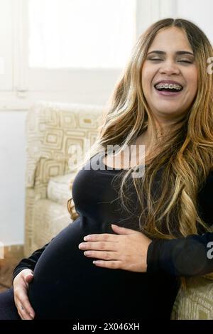Schwangere Frau lächelt in die Kamera, während eine ihrer Hände ihren Bauch berührt. Sie trägt ein schwarzes Kleid, sie ist in ihrem Haus. Stockfoto