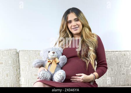 Schwangere Frau lächelt vor der Kamera, sie hat Zahnspangen an den Zähnen. Sie sitzt auf einem sehr bequemen Sofa und hält einen Teddybären in der Hand. Stockfoto