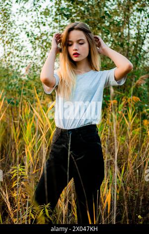 Schönes junges Mädchen, das ein leeres graues T-Shirt und eine schwarze Jeans trägt, posiert im frühen warmen Herbst vor hohem grünen und gelben Gras. Stockfoto