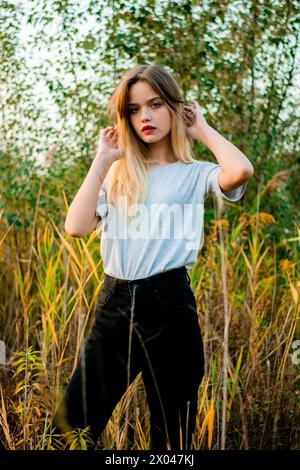 Schönes junges Mädchen, das ein leeres graues T-Shirt und eine schwarze Jeans trägt, posiert im frühen warmen Herbst vor hohem grünen und gelben Gras. Stockfoto