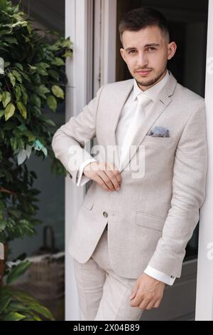 Porträt eines stilvollen Mannes, der auf der Terrasse in der Nähe des Fensters steht und vor dem Fotografen posiert. Hübsch gewachsen in hellem Anzug Stockfoto