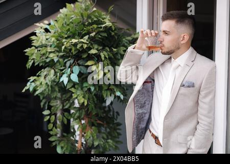 Porträt des jungen und gutaussehenden Bräutigams im hellen Anzug, der ein Glas Whiskey und Getränk hält. Seitenansicht. Platz für Text Stockfoto