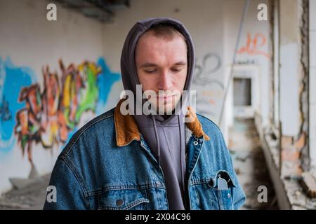 Porträt eines hübschen jungen Mannes in einer Jeansjacke und einer grauen Kapuze posiert vor dem Hintergrund eines zerstörten und verlassenen Gebäudes mit Graffiti. Stockfoto