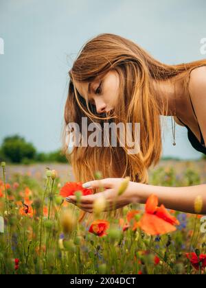 Ein wunderschönes Mädchen in einem schwarzen Abendkleid, das sich beugt, um den Duft einer Blume in einem Mohnfeld zu riechen Stockfoto