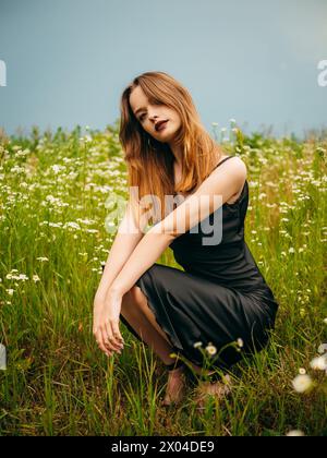 Schönes Mädchen in einem schwarzen Abendkleid posiert an einem bewölkten Sommertag vor einem Gänseblümchenfeld. Porträt eines weiblichen Modells im Freien. Stockfoto