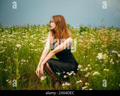 Schönes Mädchen in einem schwarzen Abendkleid posiert an einem bewölkten Sommertag vor einem Gänseblümchenfeld. Porträt eines weiblichen Modells im Freien. Stockfoto