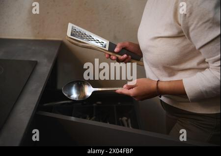 Nahaufnahme-Frau, die Küchenutensilien hält, während sie zu Hause das Abendessen kocht. Eine rostfreie Käsereibe, Löffel und Holzlöffel in den Händen der Hausfrau. Kitchenw Stockfoto