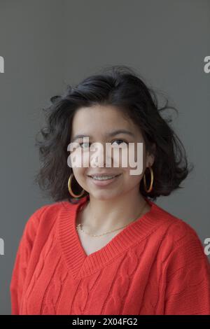 Vertikales Porträt mit grauem Hintergrund eines jungen Mädchens mit mittlerem Haar, goldenen Creolen, Zahnspangen in den Zähnen und einem roten Strickpullover. Lächeln, h Stockfoto