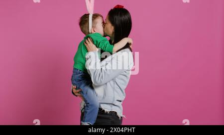 Niedliche Mutter und Kind zeigen einander Liebe im Studio, Mama umarmt und küsst ihren kleinen Kleinkinder-Sohn mit Hasenohren. Entzückende kleine Familie, die zusammen lacht und Spaß hat. Kamera B.. Stockfoto