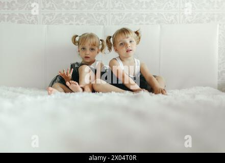 Zwei junge Mädchen sitzen nebeneinander auf einem weißen Bett und schauen sich etwas aus der Kamera an. Sie tragen legere Kleidung und wirken vertieft in Whata Stockfoto