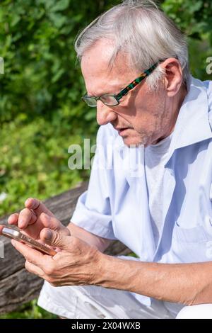 Ein älterer Mann konzentriert sich mit gemusterter Brille auf sein Smartphone in einem sonnigen, grünen Park. Das Bild erinnert an Themen der Technik in der modernen R Stockfoto