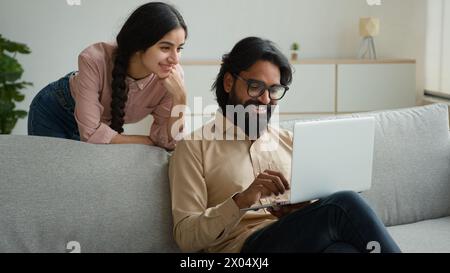 Indischer muslimischer Mann, Mann mit Laptop, reden mit Frau dahinter. Glückliche arabische ethnische Paare diskutieren über Internet-Bestellungen Kauf wählen Immobilien Stockfoto