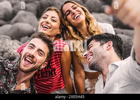 Eine fröhliche Gruppe junger Erwachsener macht gemeinsam ein verspieltes Selfie gegen graue Felsen und teilt einen glücklichen Moment des Lachens und der Verbindung in einer ungezwungenen Outdoor-Umgebung Stockfoto