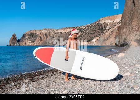 Ein Mann mit einem Weihnachtsmann-Hut trägt ein Suppe-Brett vor dem Hintergrund von Felsen, Meer, Strand. Das Konzept von Weihnachten, Reisen. Stockfoto