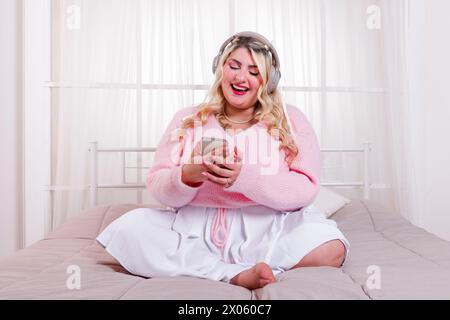 Lächelnde, barfüßige junge Frau in stilvoller Kleidung, die auf dem Bett sitzt, während sie coole Musik mit dem Headset genießt und im hellen Bett auf den Smartphone-Bildschirm blickt Stockfoto
