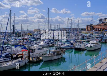 Ramsgate Royal Harbour voller verschiedener Boote und Yachten, unter einem blauen Himmel mit flauschigen weißen Wolken. Leute, die auf dem Dock laufen. Stockfoto