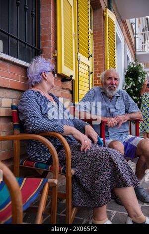 Ein älteres Paar sitzt auf einem bunten Stuhl in der Nähe einer Ziegelmauer eines Hauses mit gelben Fensterläden. Alte Frau in Sonnenbrille und Mann genießt Entspannung während Stockfoto