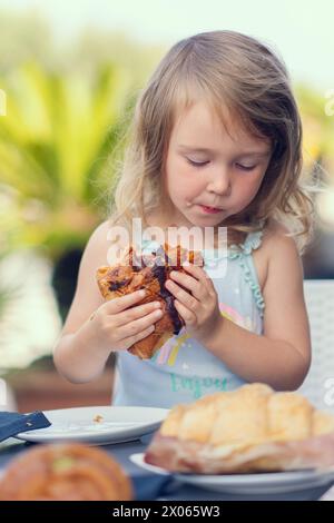 Ein hübsches kleines Mädchen isst ein Croissant mit Schokolade mit Appetit. Das Mädchen frühstückt mit einem Croissant. Das weiße Mädchen isst Gebäck. Nahaufnahme Stockfoto