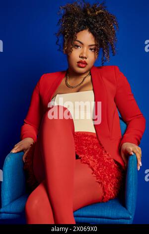 Charmante, schicke afroamerikanische Frau in elegantem rotem Anzug, die auf blauem Stuhl sitzt und in die Kamera blickt Stockfoto