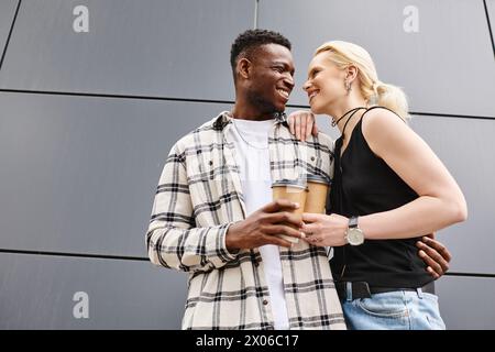 Ein fröhliches multikulturelles Paar, bestehend aus einem Mann und einer Frau, steht dicht beieinander vor einem grauen Stadtgebäude. Stockfoto