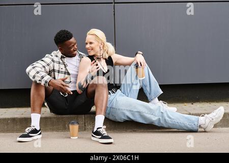 Ein Mann und eine Frau verschiedener Ethnien sehen zufrieden aus, wenn sie nebeneinander auf dem Boden in der Nähe eines grauen Gebäudes in der Stadt sitzen. Stockfoto