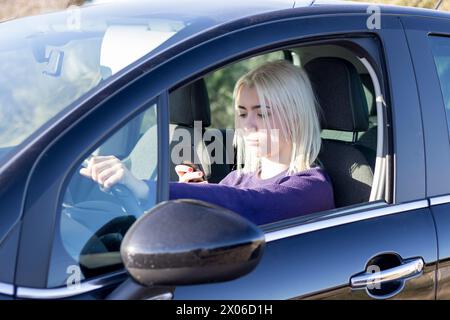 Eine lächelnde blonde Frau in einem lila Pullover, die auf ihrem Handy plaudert, während sie auf dem Fahrersitz eines Autos sitzt Stockfoto