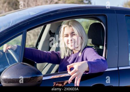 Glückliche junge Frau mit blonden Haaren, lächelnd und mit Autoschlüsseln, Sonnenbrille und lila Pullover, im Auto sitzend, Fahrersitz, Tageslicht Stockfoto