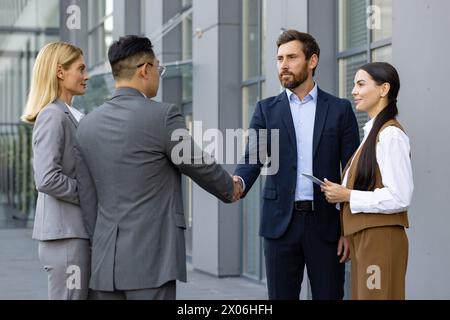 Zwei Geschäftsleute stehen vor einem Bürogebäude zusammen mit weiblichen Assistentinnen, schütteln die Hand, grüßen einander beim Treffen, vereinbaren Zusammenarbeit. Stockfoto