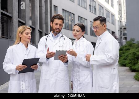 Eine Gruppe junger, interrassischer Ärzte und Wissenschaftler steht vor einem Krankenhaus in weißen Mänteln und diskutiert Daten und Forschung auf einem Tablet. Stockfoto