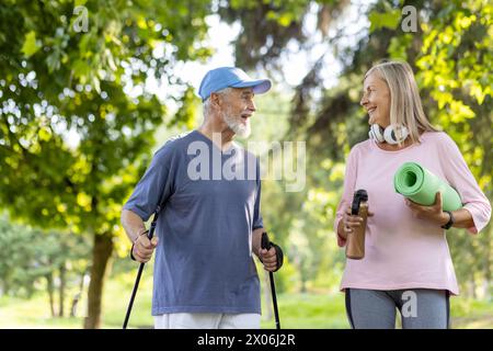 Lächelndes Paar, grauhaariger Mann und Frau, die im Park Sport treiben. Stehend mit Wanderstöcken, Matte und Wasser, miteinander reden. Stockfoto