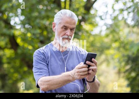 Foto eines älteren grauhaarigen Sportmannes, der im Park steht, Kopfhörer trägt und ein Mobiltelefon benutzt. Stockfoto