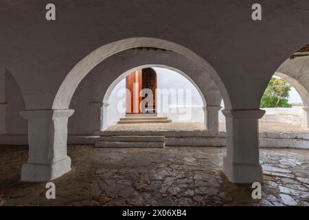 Das ruhige Ambiente des überdachten Innenhof der Kirche Puig de Missa in Santa Eulalia, Ibiza, mit seinen scharfen weißen Bögen, die einen Blick auf die Holztür bieten Stockfoto