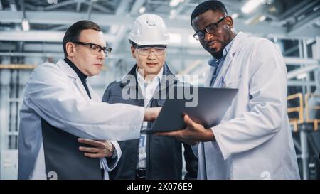 Team aus verschiedenen professionellen Ingenieuren in der Schwerindustrie, die Sicherheitsuniform und Schutzhüte tragen, die an einem Laptop arbeiten. Techniker und Mitarbeiter, die bei einer Besprechung in einer Fabrik sprechen. Stockfoto