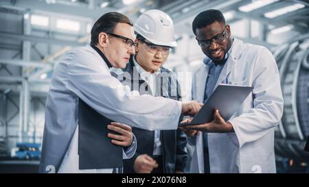 Team aus verschiedenen professionellen Ingenieuren in der Schwerindustrie, die Sicherheitsuniform und Schutzhüte tragen, die an einem Laptop arbeiten. Techniker und Mitarbeiter, die bei einer Besprechung in einer Fabrik sprechen. Stockfoto