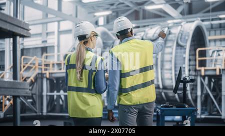 Zwei verschiedene professionelle Ingenieure in der Schwerindustrie tragen Sicherheitsuniform und Schutzhüte, die an einem Laptop arbeiten. Afroamerikanische Technikerin und weibliche Mitarbeiterin bei einem Meeting in einer Fabrik. Stockfoto
