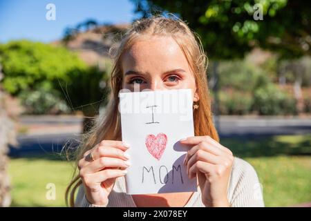 Eine Frau hält eine Karte, auf der steht: "Ich liebe dich Mom". Sie lächelt und sieht in die Kamera. Stockfoto