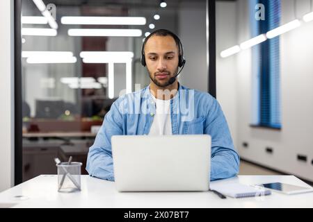Ernsthafter und konzentrierter junger hispanischer Mann, der im Studium am Laptop im Büro mit Headset arbeitet. Stockfoto