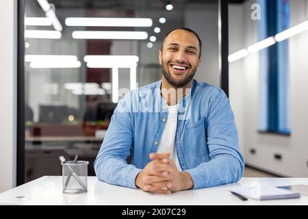 Fröhlicher indischer Mann in einem Business-Casual-Outfit, der an seinem Schreibtisch in einem modernen Büro sitzt und die Kamera anlächelt. Stockfoto