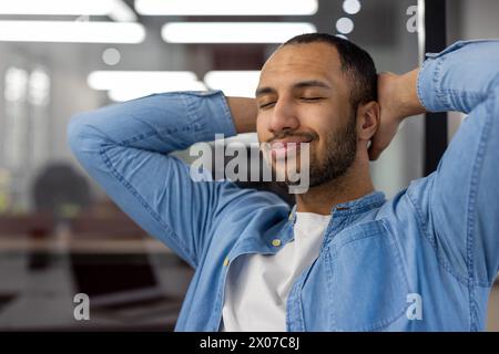 Ein junger Indianer in Denim-Kleidung, der mit geschlossenen Augen lächelt und sich in einem modernen Büroumfeld entspannt, der Ruhe und Zufriedenheit ausstrahlt. Stockfoto