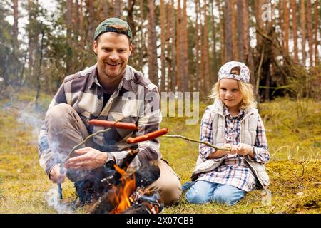 Vater und Tochter verbringen Zeit miteinander und braten Würstchen am Lagerfeuer, während sie im Wald campen. Bonding-Aktivitäten Stockfoto
